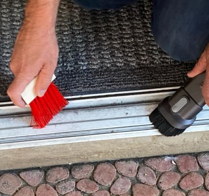 Close-up of hands cleaning a window track with a red brush and vacuum attachment. Detailing home maintenance for window cleaning.